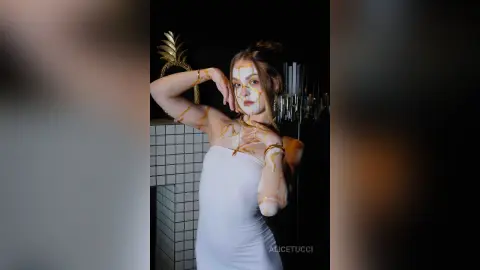 AliceTucci poses with gold liquid dripping down her face, neck, and chest while wearing a white strapless dress against a dark background.