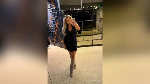 Stylish woman in a black romper and grey tall boots taking a mirror selfie in a modern building hallway.