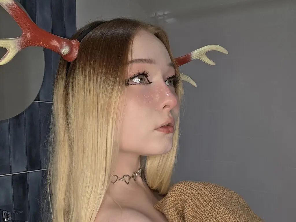 Blonde woman wearing reindeer antlers and a gold heart choker in a modern bathroom setting.