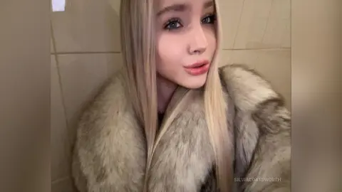 Blonde woman in a luxurious faux fur coat posing indoors with a soft, elegant mood.