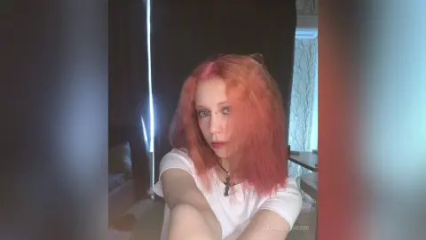 Young woman with pink hair wearing a white t-shirt and silver cross necklace in a dimly lit room.