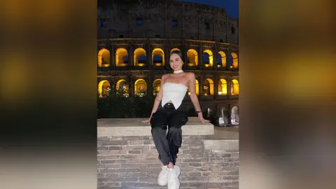 Smiling woman in a white halter top and black trousers posing in front of the illuminated Roman Colosseum at night.