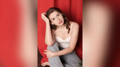 Young woman in a white tank top and jeans posing against a red backdrop in a moody studio setting.
