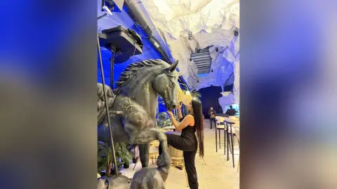 Woman in black outfit standing next to a large horse statue in a modern blue-lit lounge setting.