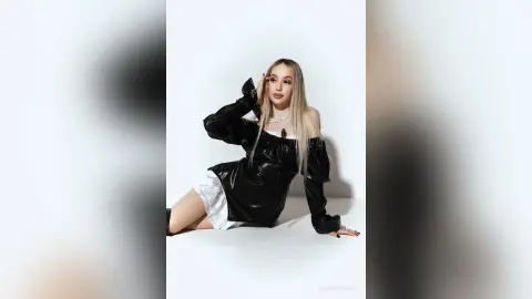 MarryDavies poses in a black leather off-the-shoulder top and white shorts, running her hand through her long blonde hair against a white background.