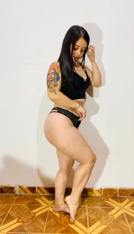 Young woman with arm tattoos wearing black lace lingerie posing against a white wall in a bright room.