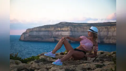 AnaMathers sits on a rocky cliffside overlooking the ocean, wearing a pink off-the-shoulder top, black panties, and a white hat.