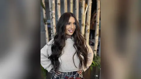 Lexi smiles with her long wavy dark hair while wearing a cozy grey sweater and plaid skirt.
