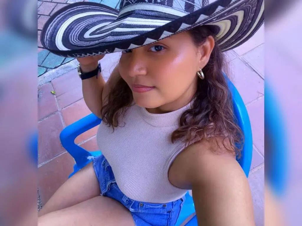 Smiling woman in a white ribbed tank top and denim shorts wearing a wide-brimmed patterned sun hat outdoors.