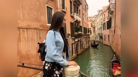 LucyMartin stands on a balcony overlooking a narrow Venice canal, wearing a light blue denim jacket and a black floral skirt.