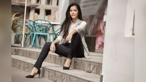 JessieLiu sits on stone steps wearing a white top, light grey blazer, and black trousers with black high heels.