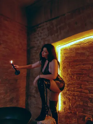 Moody portrait of a woman in a black outfit holding a candle in a rustic brick room with warm lighting.
