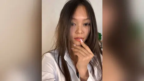 Young woman with long brown hair wearing a white button-down shirt in a bright, minimalist indoor setting.