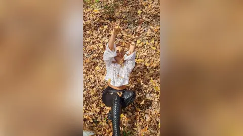 Young woman in a white crochet top sitting in autumn leaves with arms raised in a joyful forest setting.