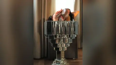 MissyJolie lies on her back with her legs raised and feet resting on a silver chrome stand.