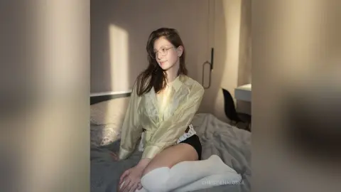 Young woman in a pale yellow silk blouse and black shorts sitting on a bed in soft, natural sunlight.
