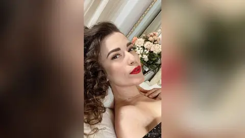 MontseFerrera poses with a direct gaze, wearing red lipstick and a black strapless top with her long, curly brown hair falling over one shoulder.