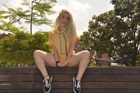 Smiling blonde woman in a yellow hoodie sitting on a wooden bench outdoors in a sunny park setting.