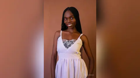 Smiling Black woman in a white sleeveless dress posing against a warm brown background.