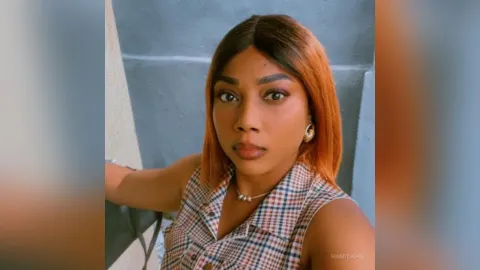 MandyJohn — MandyJohn looks into the camera wearing a plaid sleeveless shirt and gold earrings.