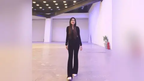 Elegant woman in a black long-sleeve jumpsuit standing in a minimalist, brightly lit modern studio space.