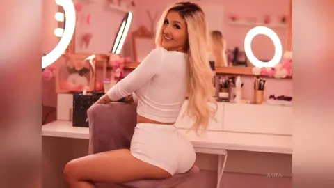 Anita poses sitting on a grey chair in a white long-sleeve crop top and matching white shorts, showing her blonde hair and bare legs.