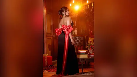 Elegant woman in a black strapless gown with large red bows posing in a warm, moody festive room.