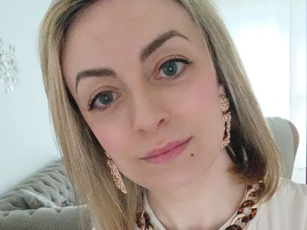 Blonde woman with gold statement earrings smiling in a bright, casual indoor setting.