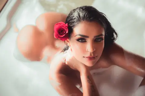Emilycurtiis lies naked in a bubble bath with a red rose in her dark hair, looking up at the camera.