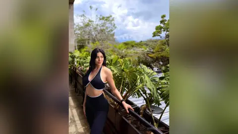 Athletic woman in a black halter sports top and leggings posing on a tropical balcony under a cloudy sky.