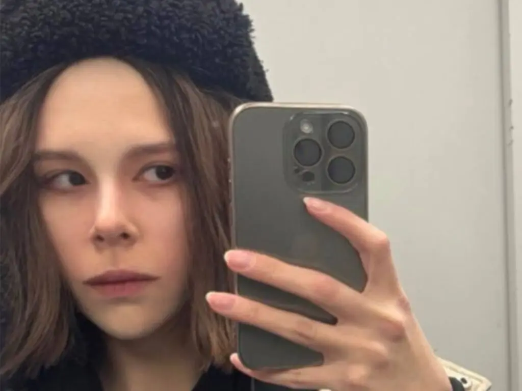 Young woman with long brown hair wearing a black fuzzy hat taking a moody mirror selfie with a smartphone.