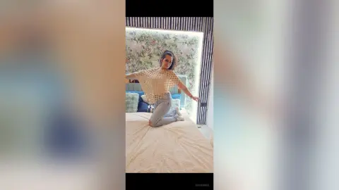 Smiling woman in a beige crochet top and light blue jeans sitting on a bed in a bright, floral bedroom.