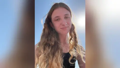Smiling young woman with long wavy hair in a black tank top outdoors under a bright sunny sky.