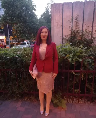Woman with purple hair in a red blazer and champagne lace dress standing by a garden fence at dusk.