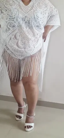 Plus size woman in a white lace bohemian fringe tunic and strappy sandals standing against a neutral wall.