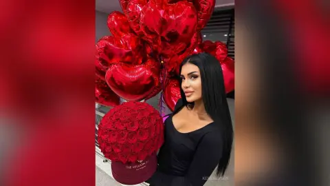 Beautiful woman in a black dress holding red roses near large red heart balloons for a romantic celebration.