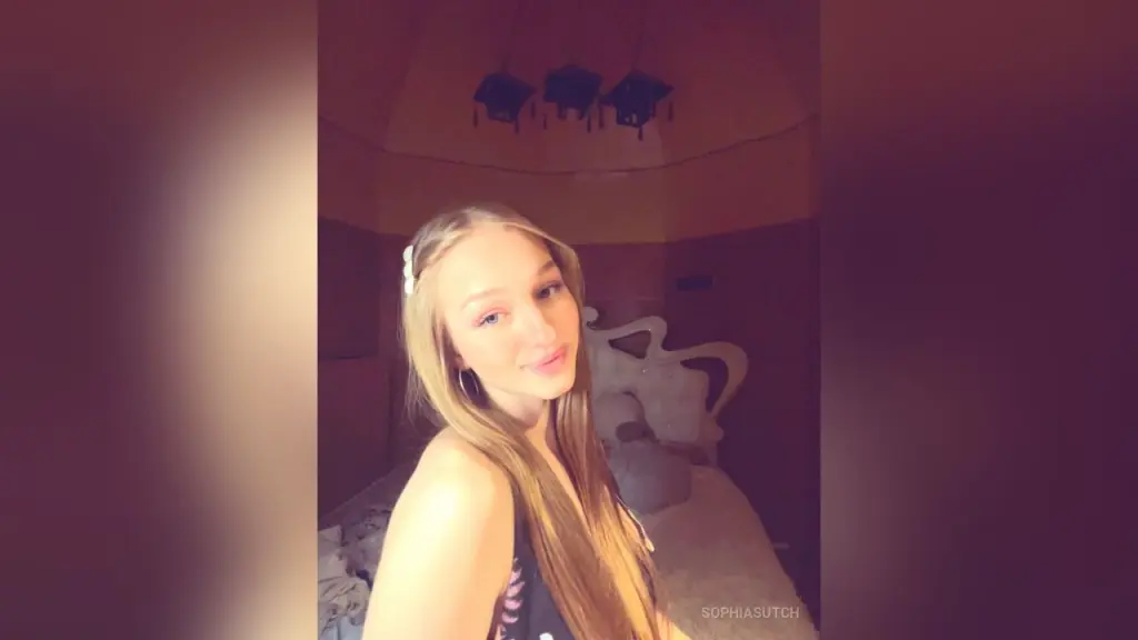 Blonde woman in a black tank top taking a selfie in a bright marble bathroom.