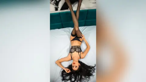 OliviaLarson lies on a white bed wearing black lace lingerie and black stockings, spreading her long black hair out behind her head.