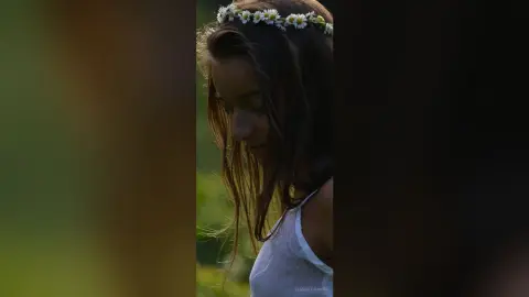 Dreamy portrait of a young woman wearing a white tank top and daisy flower crown in a sunny meadow.