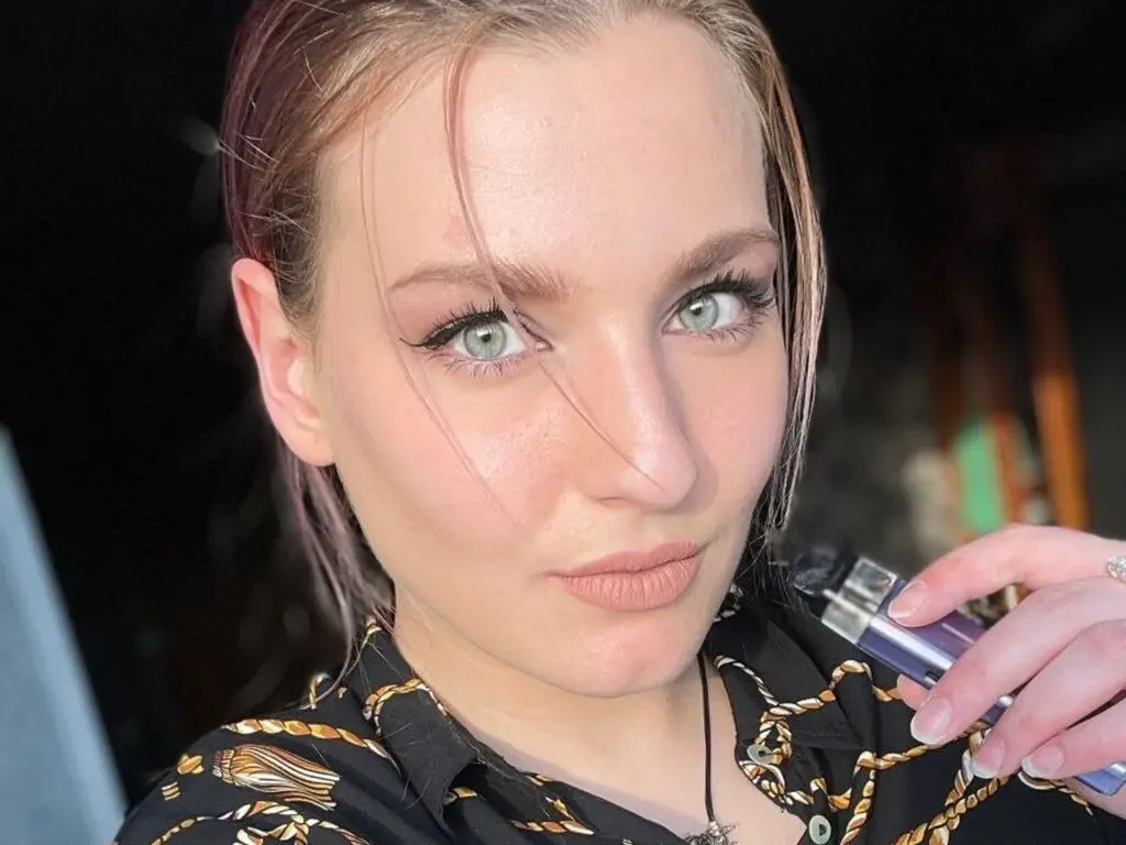 CristaStwell looks into the camera with light eyes and wet-look hair, wearing a black patterned shirt while holding a purple vape pen.