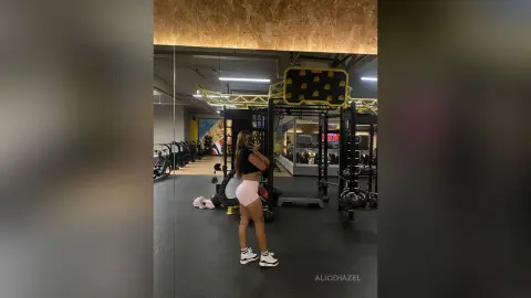 Fit woman in black crop top and pink shorts taking a selfie in a modern gym setting.
