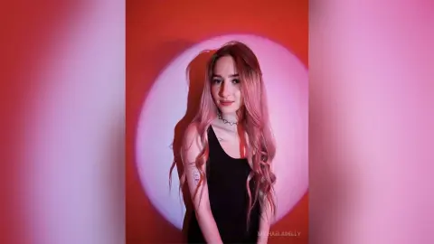 MichaelaDelly poses against a red wall wearing a black tank top and a silver choker necklace.