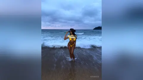 Stylish woman in a yellow bikini and black hat standing on a moody, cloudy beach at dusk.