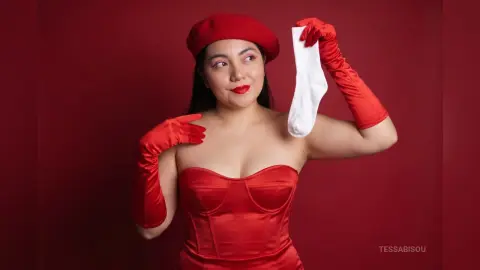 Elegant woman in red satin corset and beret holding a white sock against a deep red background.
