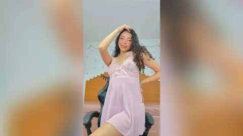 Smiling young woman in a pink lace slip dress posing in a bright bedroom setting.