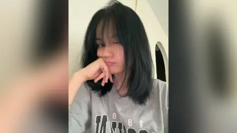 Young woman with long dark hair wearing a grey graphic t-shirt in a bright indoor setting with a relaxed mood.