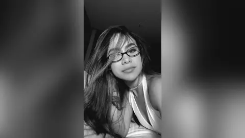 Young woman with long hair and glasses wearing a white tank top in a moody black and white portrait.