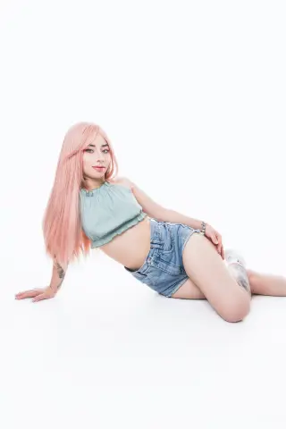 Young woman with long pink hair in a sage green crop top and denim shorts posing against a white background.