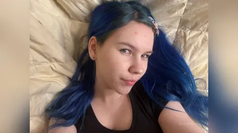 DeidreSarin has long blue hair and is wearing a black tank top while lying on a tan bedsheet.