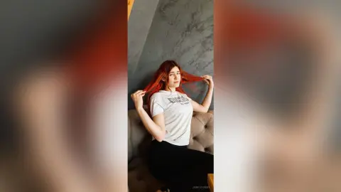 Red-haired woman in a white t-shirt posing thoughtfully on a grey sofa in a modern minimalist room.